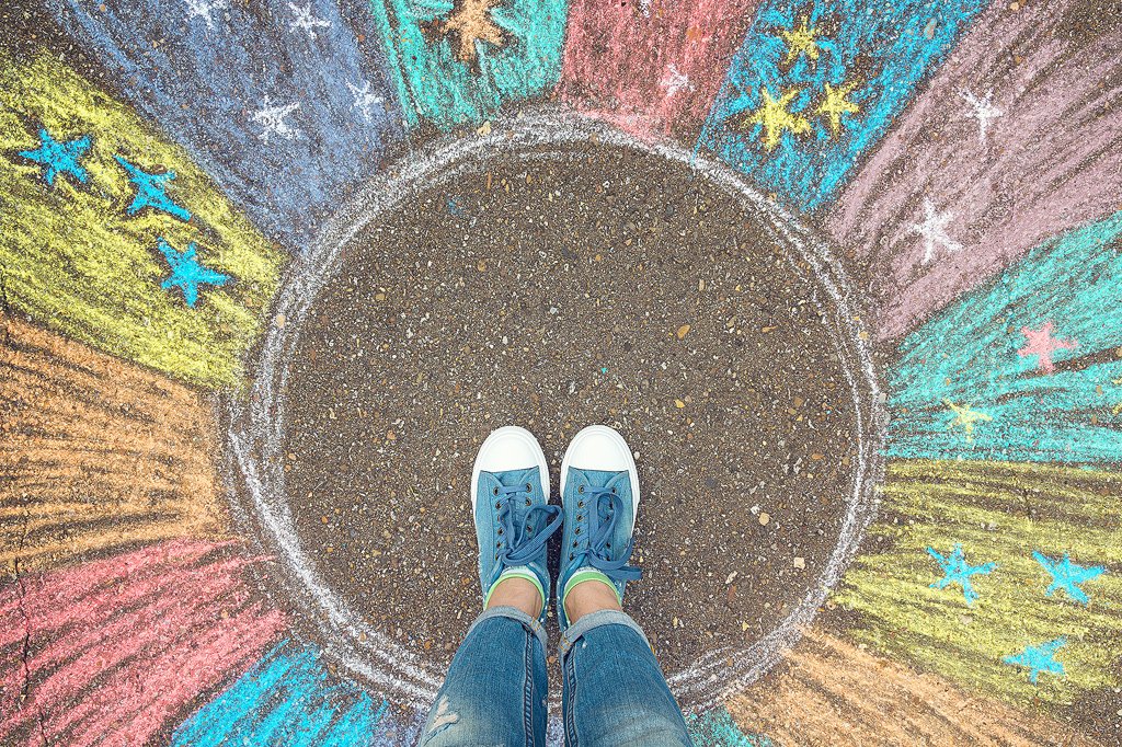 Eine illustrative Aufnahme der Komfortzone. Füße, die auf einem unmarkierten Stück Straße stehen, umgeben von spritzenden Farben und Sternen, die verdeutlichen, wie magisch es sein kann, aus seiner Komfortzone herauszutreten.